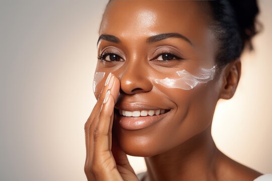 Close-up of young African American woman touching her face to apply moisturizer. Smiling face of beautiful colored lady with daily cream, facial cosmetics. Skin care. Monochrome background, copy space