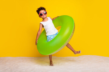 Full length photo of funky positive little boy dressed white shirt walking beach inflatable ring...