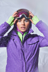 Fototapeta na wymiar attractive woman in balaclava and winter jacket wearing ski googles on grey backdrop, snowy day