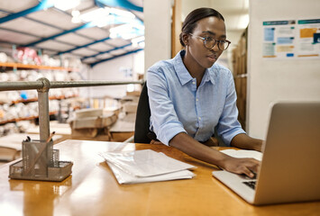 Young African woman sitting at her warehouse desk working on a laptop