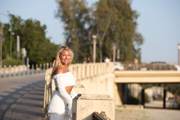 Beautiful young blonde woman from the United States is sightseeing in Seville, Spain. The girl is leaning over the railing. In the background trees and a bridge over the river guadalquivir.