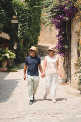 Fototapeta na wymiar Retired couple wearing hats and sunglasses enjoy strolling through the picturesque village of Peratallada, Catalonia. On a sunny day, in a Mediterranean heritage destination.
