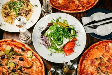 A feta cheese Greek salad surrounded y different pizzas on a wooden table.
