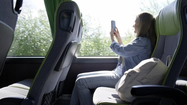 A blonde woman is filming on a smartphone in a tourist bus, sitting and looking out the window
