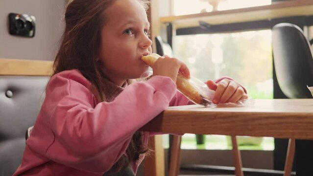 Little girl wearing pink sweatshirt sitting at table in cafe schoolgirl after classes eating bun sitting by window looking away at something lunch after school.