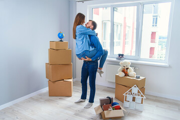 Front view of pretty smiling girl which holds by her handsome young boyfriend in the middle of their new flat full of cardboard boxes.