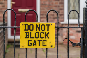 Yellow do not block gate sign with a house with a red door in the background, information and...