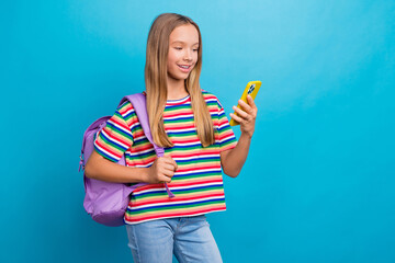 Photo of good mood schoolgirl dressed striped t-shirt hold rucksack read notification on smartphone isolated on blue color background