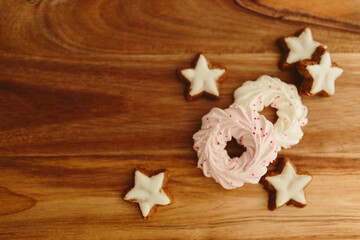 Fototapeta premium New Year background for winter cards. Gingerbread cookies in the shape of stars and pink meringue cookies lie on a brown background with Christmas lights. Delicious sweet pastries on a wood table