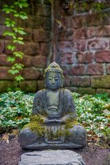a grey stone buddha with moss in front of an old red brick wall and ivy