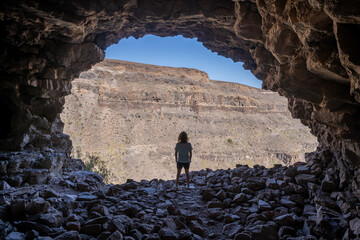 Rear view of a woman standing in the access of a large cave looking outside in the archaeological...