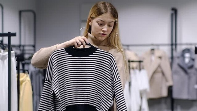 A girl tries on a striped sweater in a store. A young woman chooses clothes in a store while shopping. Seasonal holiday discounts on purchases. Black Friday