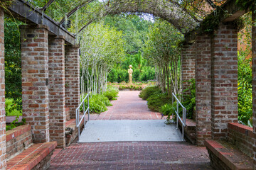 A long red brick footpath with a metal awning covered with lush green plants and trees with a gold statue at the end of the footpath at New Orleans Botanical Garden in New Orleans Louisiana USA