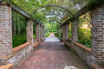 A long red brick footpath with a metal awning covered with lush green plants and trees with a gold statue at the end of the footpath at New Orleans Botanical Garden in New Orleans Louisiana USA