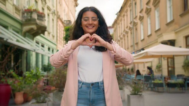 Smiling happy young teen Indian Arabian woman female lady girl making heart shape hands to camera laughing positive gesture love like emoji support care sign symbol standing outside town city street