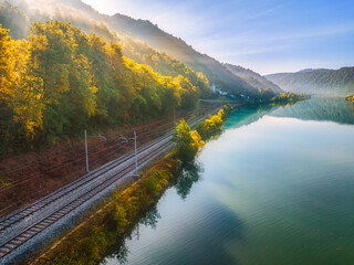 Aerial view of railroad near river in alpine mountains in fog at sunrise in autumn. Top view of rural railway station, lake, reflection in water, orange trees in fall. Railway station in Slovenia