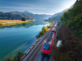 Aerial view of red modern high speed train moving near river in alpine mountains in fog at sunrise in autumn. Top view of train, railroad, lake, reflection, trees in fall. Railway station in Slovenia
