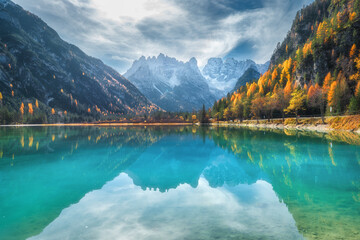 Mountain lake with reflection at sunny autumn day in Dolomites, Italy. Beautiful landscape with azure water, orange trees, snowy mountains, blue sky with clouds in fall. Snow covered rocks. Nature - obrazy, fototapety, plakaty