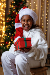 Cheerful dark-skinned teenage girl in a Santa hat holds a gift box in her hands, near the Christmas...