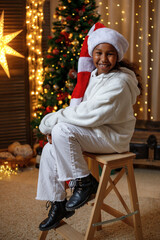 Cheerful dark-skinned teenage girl wearing a Santa hat, near the Christmas tree at home. Christmas...