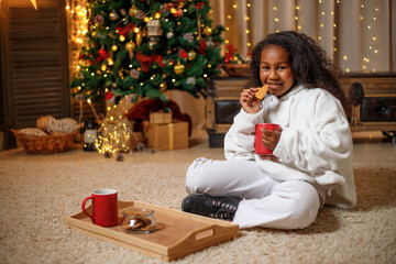 Cheerful dark-skinned teenage girl holding a red mug with cocoa in her hands, near the Christmas...