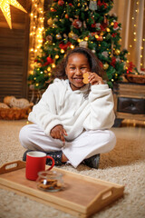 Cheerful dark-skinned teenage girl holding a red mug with cocoa in her hands, near the Christmas...