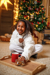 Cheerful dark-skinned teenage girl holding a red mug with cocoa in her hands, near the Christmas...