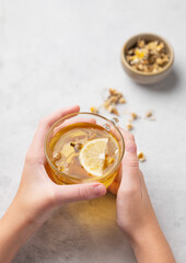 Hands holding a glass cup with chamomile herbal tea on a light background with dry flowers. The concept of a healthy drink for health and immunity.