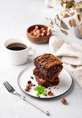 A stack of chocolate brownies with mint, hazelnuts and salted caramel on a white plate on a light background with cup of coffee and bowl with nuts.