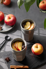 Homemade apple punch or cider with apples and cinnamon in gray cups on a dark background with fresh fruits, spices and shadows.