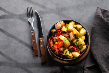 Fried pork meat with potatoes, onions and peppers. National homemade Georgian dish ojakhuri in a black frying pan on a dark background with shadows.