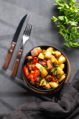 Fried pork meat with potatoes, onions and peppers. National homemade Georgian dish ojakhuri in a black frying pan on a dark background with fresh herb and shadows.