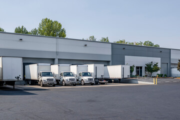 Middle duty day cab white rigs semi trucks with box trailers standing in row with dry van semi trailers in warehouse docks loading cargo for the next delivery