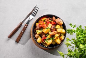 Fried pork meat with potatoes, onions and peppers. National homemade Georgian dish ojakhuri in a black bowl on a light background with fresh herb.