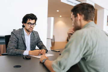 Selective focus of broker advisor in eyeglasses and business suit consulting to young man client sitting at desk. Professional male insurer talking consumer guy about insurance benefits at office. - obrazy, fototapety, plakaty