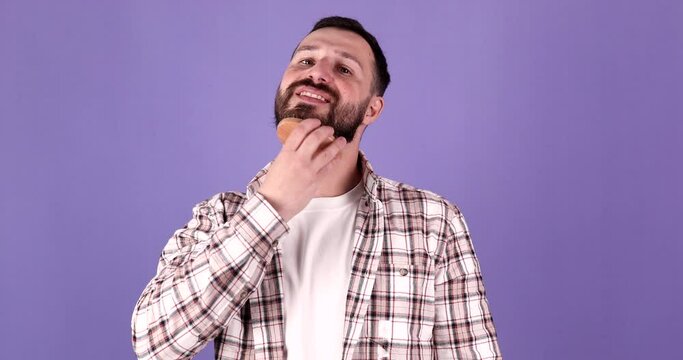 Handsome young bearded guy, brutal man with a beard hair is combing the beard on face with a comb, brush on purple background. Male personal self care, products, accessory.