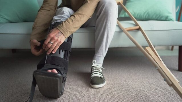 Young man puts on ankle brace while sitting on the couch at home after a serious injury.