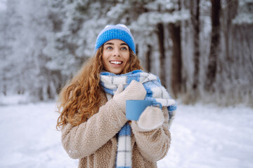 Obraz premium Happy woman holding a thermos and drinking tea in the winter forest. Vacation, relaxation, travel, lifestyle concept. Happy winter days.