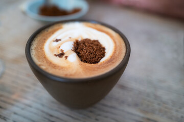 Dark brown granulated sugar before stirring on the froth of a flat white coffee on a wooden table in a mug with no handle.