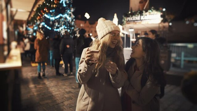 Young women outdoor spend time together at Christmas New Year holidays at illuminated background of decorated fairy at town square. Best friends tourists in warm knitted clothes enjoy winter vacation