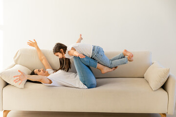 Joyful young strong woman lifting on legs arms laughing little child daughter, having fun together on cozy sofa in modern studio living room. Overjoyed small kid girl practicing acroyoga with mom. - obrazy, fototapety, plakaty