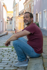 An elderly man sits on the pavement on a narrow European pedestrian street and smiles.