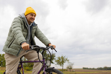 Middle aged caucasian man in warm casual clothes and yellow hat drives a bicycle in a countryside....