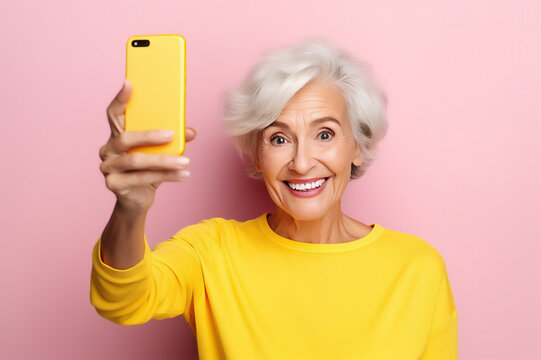 Happy 60 year old woman in yellow sweater taking selfie in studio on pink background