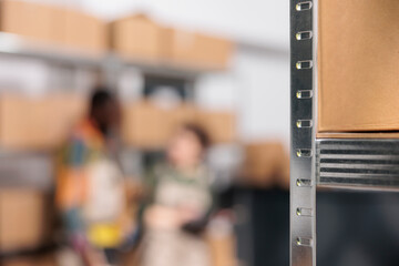 Selective focus of metallic shelves full with cardboard boxes, in background diverse workers discussing goods inventory report. Storage room team working at customers orders, preparing packages