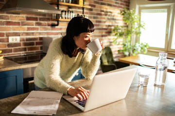Young Asian woman drinking coffee and doing financials with a laptop in the kitchen