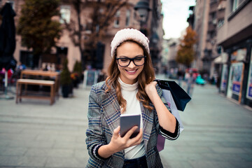 Young attractive woman using her smartphone while shopping in the city - obrazy, fototapety, plakaty