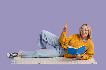 Beautiful young happy woman with book lying on lilac background and pointing at something