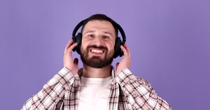 Portrait of attractive laughing bearded man wearing large wireless headphones on purple background.