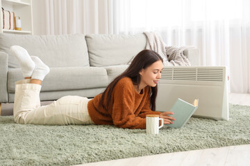 Obraz premium Pretty young woman reading book near electric heater in living room